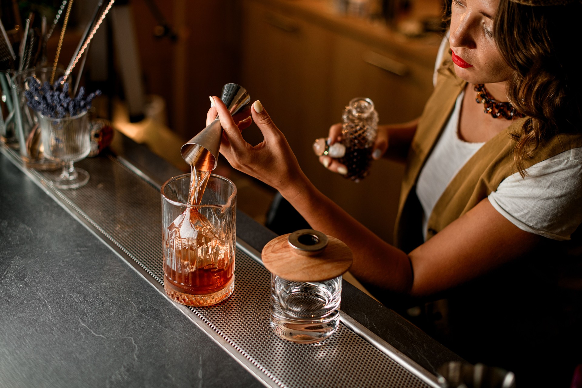 Top view of mixing glass in which bartender pours brown drink from jigger